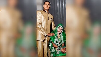 A young man in a gold sherwani standing beside a seated woman in vibrant green bridal dress and jewelry,.