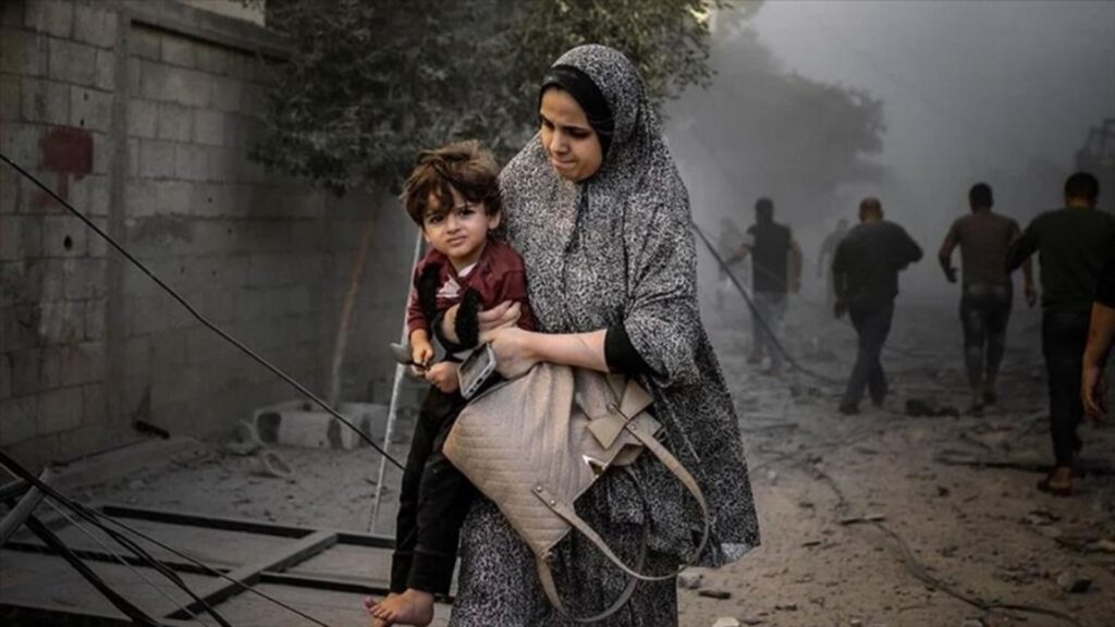 Woman carrying a child through a smoky, war-torn area with other people in the background.