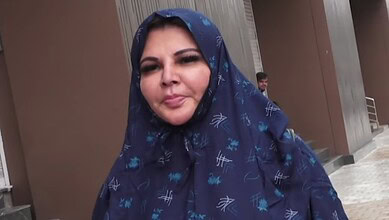 A woman wearing a blue patterned headscarf, smiling outdoors in front of a modern building.