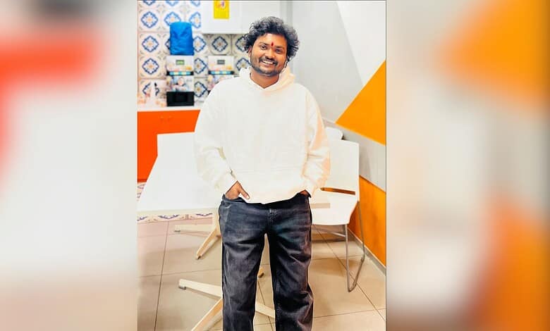 Young man with curly hair smiling in a contemporary office environment, casual attire, and relaxed pose.