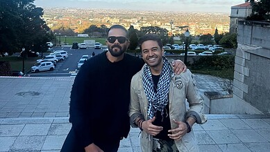 Two men smiling outdoors with a cityscape in the background during daytime.