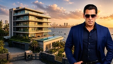 Salman Khan in a blue suit and sunglasses standing in front of a modern waterfront building at sunset.