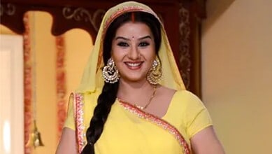 A woman in traditional yellow attire with jewelry, smiling in an indoor setting.