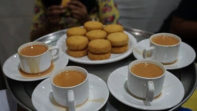 Tea cups with saucers and a plate of cookies on a tray, with a person in the background.