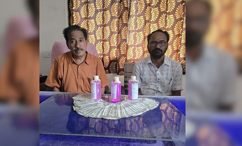 Two officials seated at a table with pink bottles and a silver cloth, curtain backdrop.