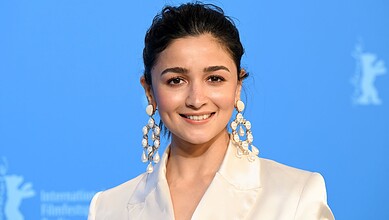 Alia Bhatt smiling at an event with a blue background and white logo.