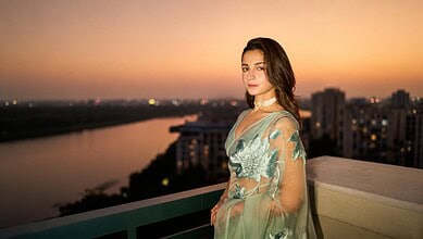 Alia Bhatt standing on balcony during sunset with cityscape in background.