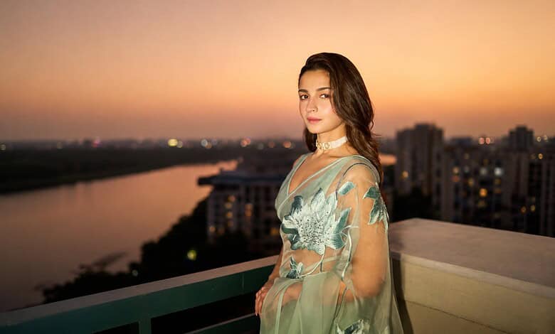 Alia Bhatt standing on balcony during sunset with cityscape in background.