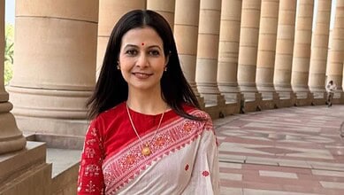 Rukmini Mallick in a red and white saree, smiling outside a grand building with columns.