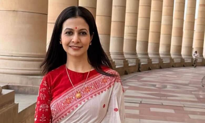 Rukmini Mallick in a red and white saree, smiling outside a grand building with columns.