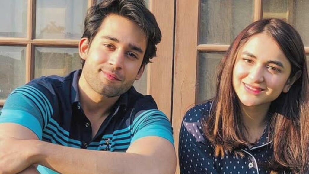Young man and woman smiling outdoors in front of a wooden window.