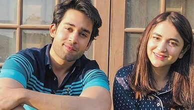 Young man and woman smiling outdoors in front of a wooden window.