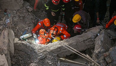 Rescue workers in orange uniforms and helmets working amidst debris after building collapse.