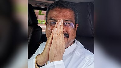 Indian man with glasses and mustache, hands pressed together in a gesture of respect inside a vehicle.