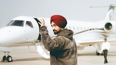 Man wearing a red turban and brown jacket waving near a private jet at an airport.