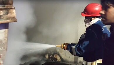 Firefighter in red helmet spraying water on a fire with smoke in the background.