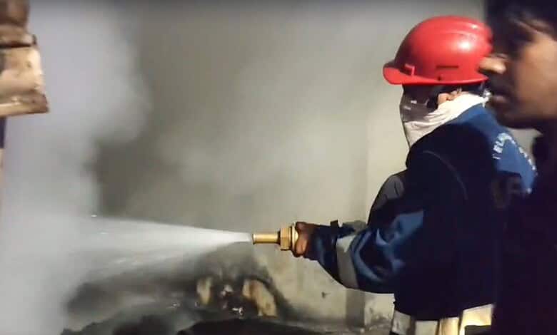 Firefighter in red helmet spraying water on a fire with smoke in the background.