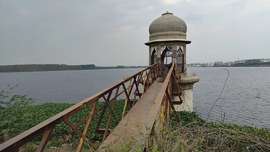 View of a small pavilion on a lake with a wooden bridge leading to it, under cloudy sky.