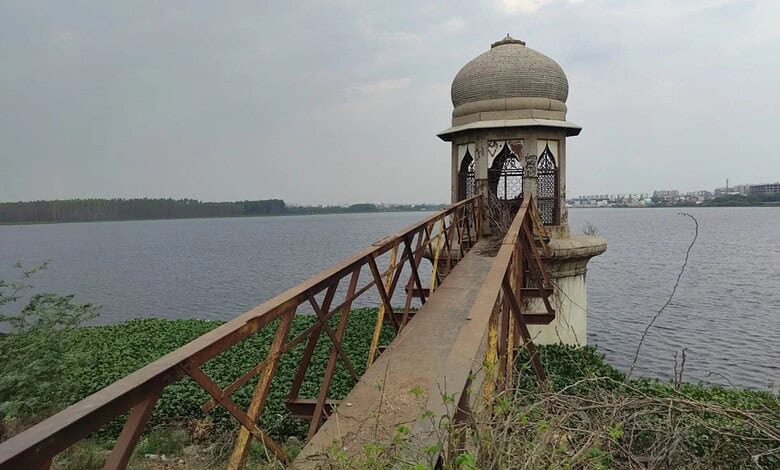 View of a small pavilion on a lake with a wooden bridge leading to it, under cloudy sky.