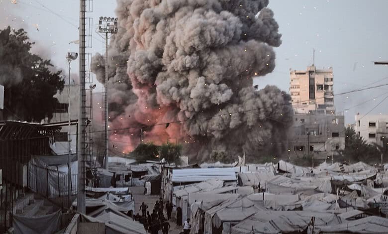 Explosion with thick black smoke rising over a cityscape with tents and buildings.