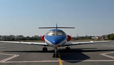 Small private jet on runway with cityscape in background, clear sky, daytime.