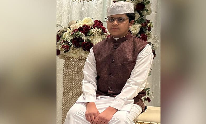 Young boy in traditional Indian attire with glasses and a cap, sitting near a floral arrangement.