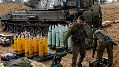 Soldiers prepare military equipment near a tank with artillery shells on the ground.