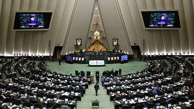 Iranian parliament session with members seated and screens displaying a speaker.