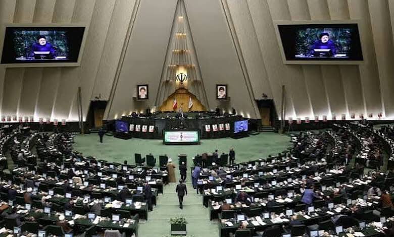 Iranian parliament session with members seated and screens displaying a speaker.