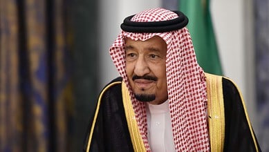 Saudi official in traditional attire at a formal event with green and gold background.