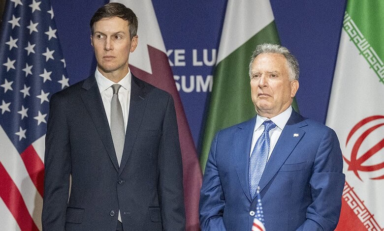 Two men in suits standing in front of flags at a formal event.