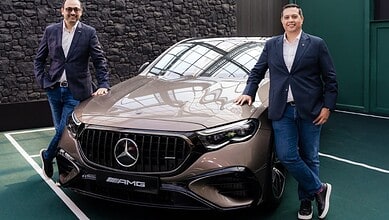 Two men in formal attire stand beside a beige Mercedes AMG E 53 Hybrid car.