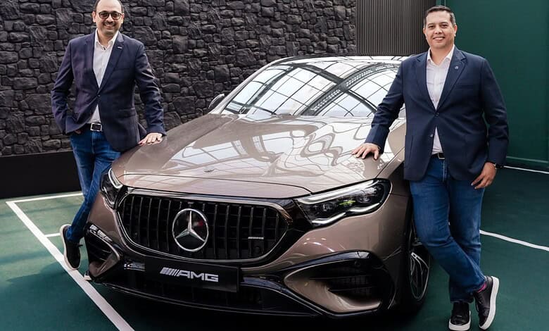 Two men in formal attire stand beside a beige Mercedes AMG E 53 Hybrid car.