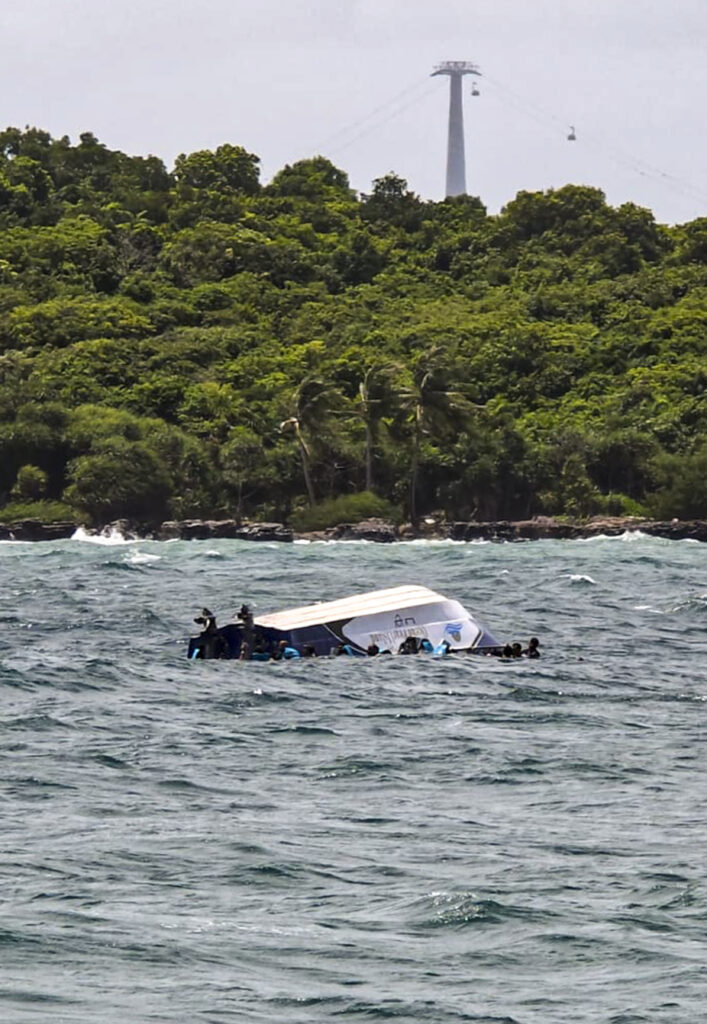Boat capsized in water near lush green shoreline with a tower in the background.