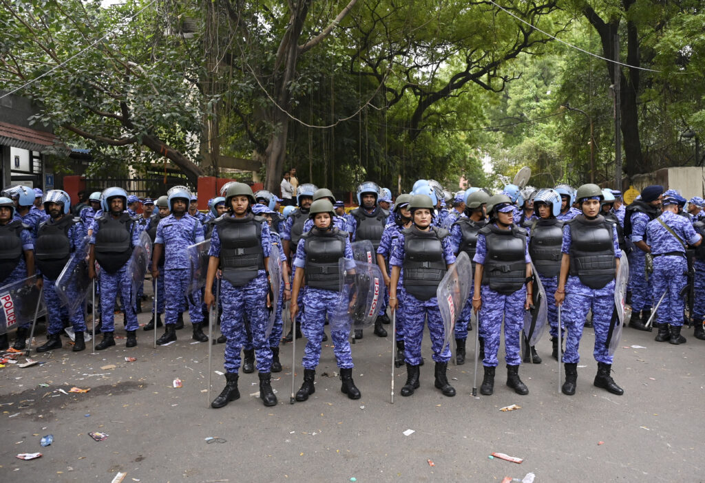 Line of police officers in riot gear standing on a street with trees in the background.