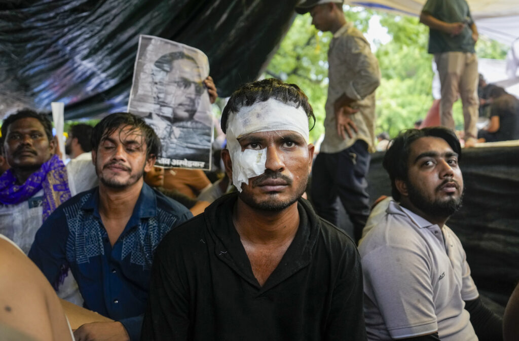 Protesters with bandaged face at a rally, holding a portrait of a leader, under a tent.