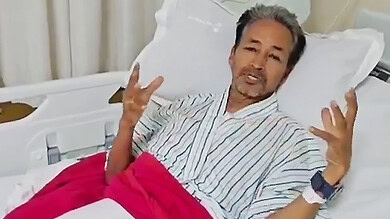 Elderly man speaking while lying in hospital bed with striped shirt and red blanket.