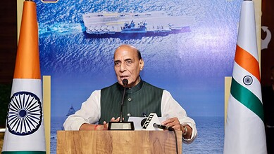 Indian official speaking at a podium with Indian flags and a ship image in the background.