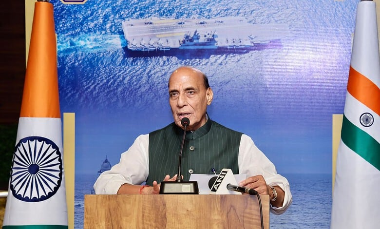Indian official speaking at a podium with Indian flags and a ship image in the background.