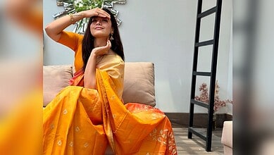 Rashmika Mandanna wearing a yellow saree, sitting on a sofa, smiling and shielding her eyes from the sun.