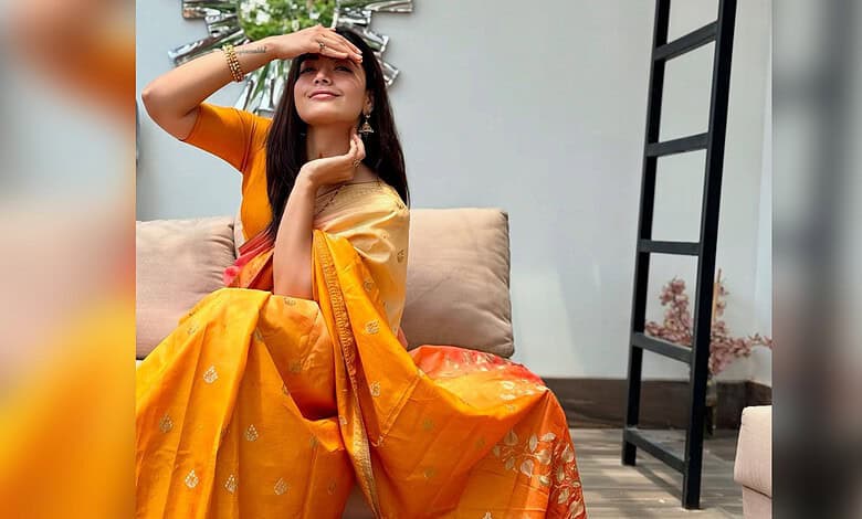Rashmika Mandanna wearing a yellow saree, sitting on a sofa, smiling and shielding her eyes from the sun.
