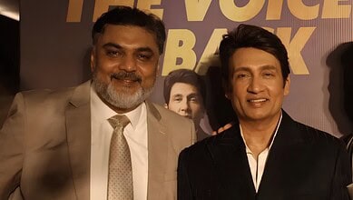 Two men in formal attire smiling at an event, with a backdrop featuring a person's face and text.