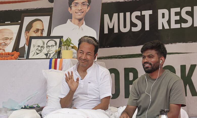 Sonam Wangchuk protesting with a smile at a rally, surrounded by portraits and banners.