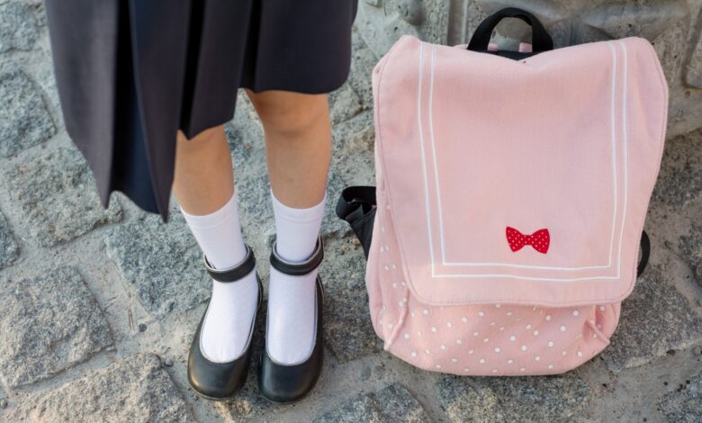 Schoolgirl's legs with white socks and black shoes next to a pink backpack on stone ground.