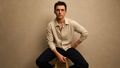 Tom Holland sitting on a stool, wearing a beige shirt and black pants, against a plain background.