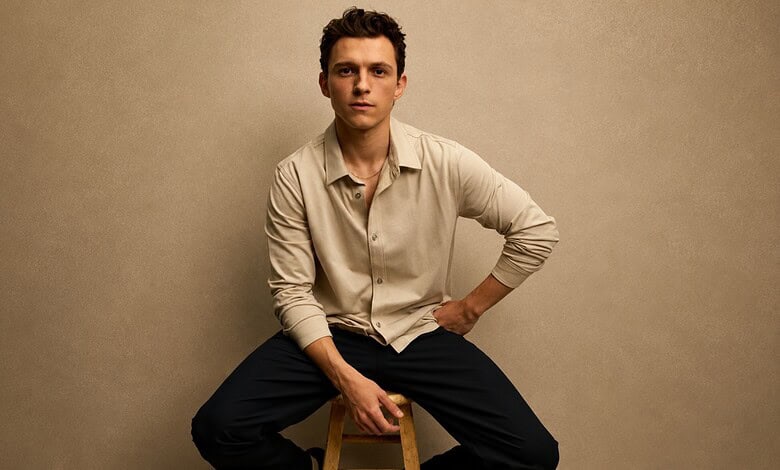 Tom Holland sitting on a stool, wearing a beige shirt and black pants, against a plain background.