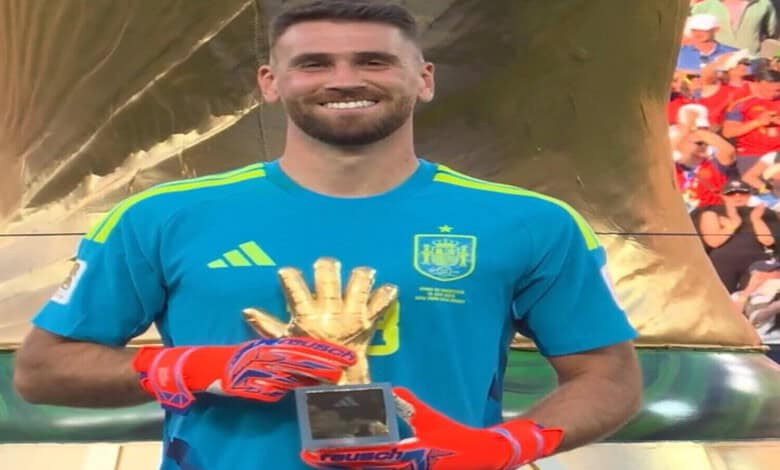 Smiling goalkeeper in blue jersey holding a trophy with gloves, celebrating victory.