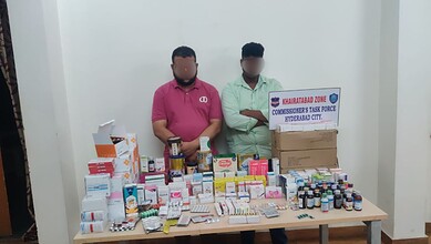 Two men with blurred faces stand behind a table of medicines in Khairatabad.