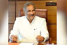 Senior man in white kurta smiling and sitting at a desk in an office setting.