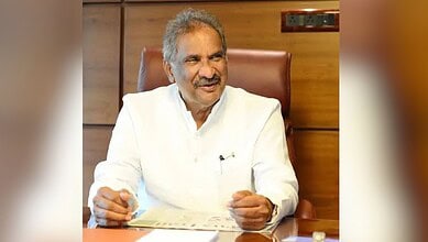 Senior man in white kurta smiling and sitting at a desk in an office setting.